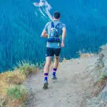 Eine Person joggt auf einem schmalen Pfad am Berghang. Die Person trägt einen Rucksack und Sportkleidung. Im Hintergrund erstreckt sich ein dichter Nadelwald, der die Hänge des Berges bedeckt.
