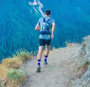 Eine Person joggt auf einem schmalen Pfad am Berghang. Die Person trägt einen Rucksack und Sportkleidung. Im Hintergrund erstreckt sich ein dichter Nadelwald, der die Hänge des Berges bedeckt.