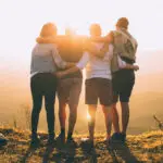 Vier Personen stehen auf einer Wiese und umarmen sich von hinten bei Sonnenuntergang, wobei die Sonne zwischen ihnen durchscheint. Sie blicken auf eine Landschaft im Hintergrund.