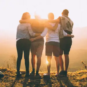 Vier Personen stehen auf einer Wiese und umarmen sich von hinten bei Sonnenuntergang, wobei die Sonne zwischen ihnen durchscheint. Sie blicken auf eine Landschaft im Hintergrund.