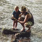 Ein Mann in Shorts und Hut kniet neben einem Kind auf einem flachen Stein in einem flachen Fluss. Beide sind barfuß und schauen sich einen großen Stein an, den der Mann hält. Das Flusswasser fließt ruhig um sie herum.