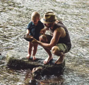Ein Mann in Shorts und Hut kniet neben einem Kind auf einem flachen Stein in einem flachen Fluss. Beide sind barfuß und schauen sich einen großen Stein an, den der Mann hält. Das Flusswasser fließt ruhig um sie herum.