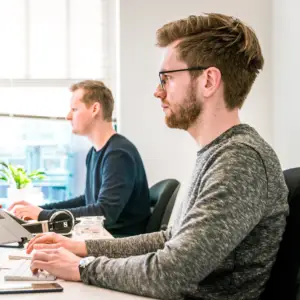 Zwei Männer sitzen in einem hellen Büro nebeneinander an Computern. Der Mann im Vordergrund hat eine Brille und tippt auf einer Tastatur. Im Hintergrund ist eine Pflanze auf dem Tisch zu sehen.