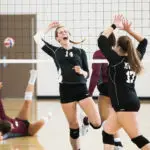 Zwei Volleyballspielerinnen in schwarzen Trikots jubeln und schlagen die Hände über dem Kopf zusammen. Eine Spielerin im weinroten Trikot liegt auf dem Boden mit ausgestrecktem Arm, während der Volleyball über das Netz fliegt. Die Szene spielt sich in einer Turnhalle ab.