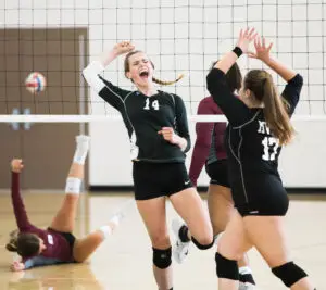 Zwei Volleyballspielerinnen in schwarzen Trikots jubeln und schlagen die Hände über dem Kopf zusammen. Eine Spielerin im weinroten Trikot liegt auf dem Boden mit ausgestrecktem Arm, während der Volleyball über das Netz fliegt. Die Szene spielt sich in einer Turnhalle ab.