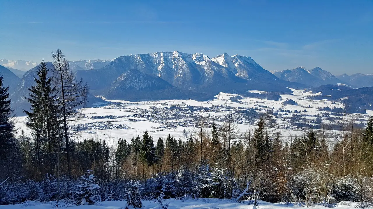 Verschneite Berglandschaft mit einem Tal im Hintergrund. Im Vordergrund stehen schneebedeckte Nadelbäume, dahinter erstreckt sich ein verschneites Tal mit vereinzelten Gebäuden. Im Hintergrund sind schroffe, schneebedeckte Berge unter einem klaren blauen Himmel zu sehen.